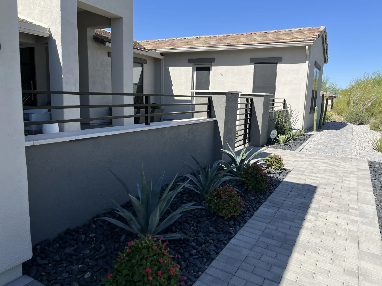 Gray stucco home with paver walkway, black river rock beds, and dark metal railing after exterior painting